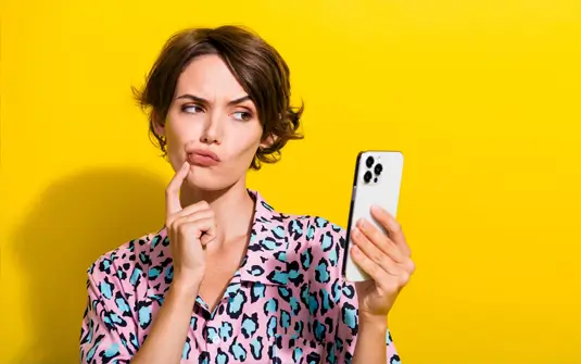 Woman unsure about purchase on mobile phone with a yellow background.